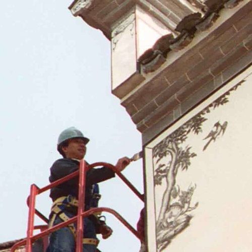 A person in a safety harness and blue helmet stands on a red scaffold, painting a detailed mural of a tree and birds on the side of a gray building.