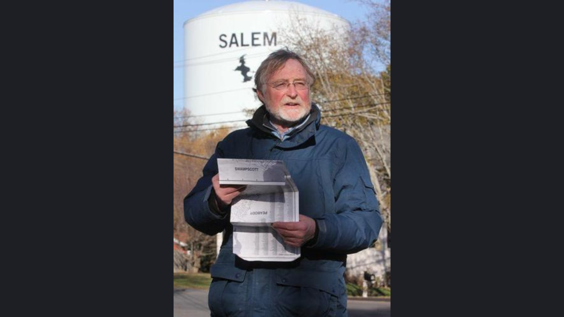 A man wearing glasses and a blue jacket stands outdoors holding documents; behind him is a large white water tower labeled “SALEM” with a witch silhouette, evoking Literary Salem. Leafless trees appear in the background under a clear sky.