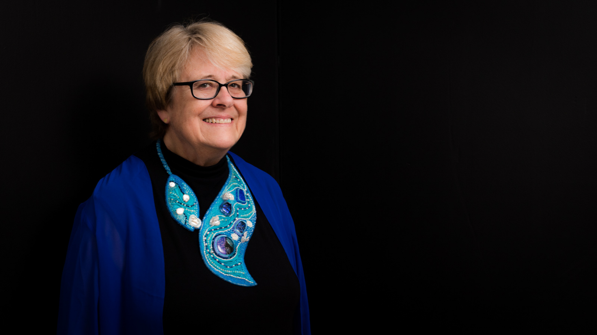 Meet the Member: Alisa Sickora, an older woman with short blond hair and glasses, smiles warmly in a black top, vivid blue shawl, and ornate blue beaded necklace against a plain black background.