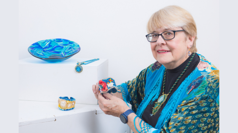 A woman with short blonde hair and glasses sits beside a white display featuring Seaside Glass—a blue glass bowl, blue necklace, and decorated cuffs. She wears a blue shawl and jewelry, holding a colorful beaded cuff created with intricate beading techniques.