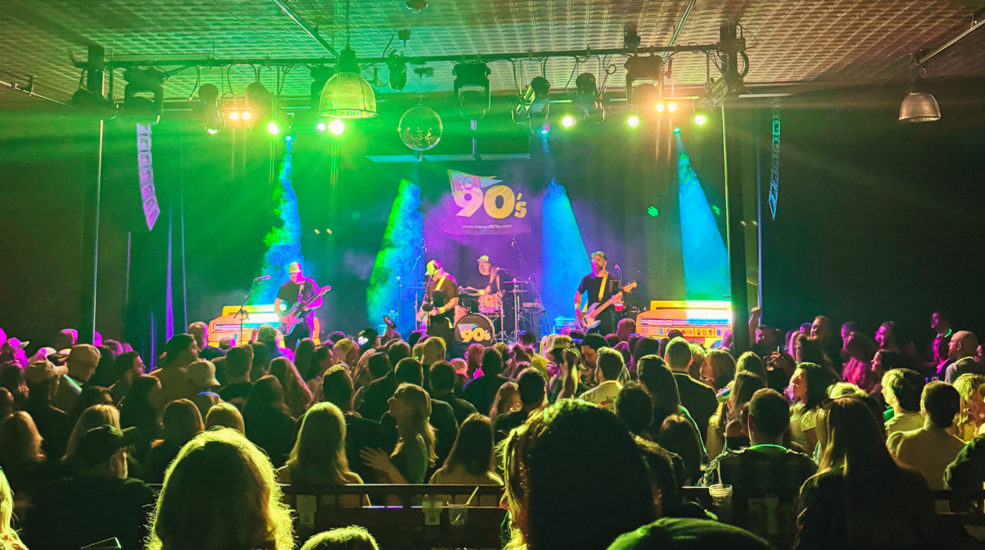 A live band, The Cut Live, performs onstage under green and yellow lights before a large crowd. A sign reading “90s” glows as audience members face the stage beneath a venue ceiling lined with lighting equipment and fixtures.