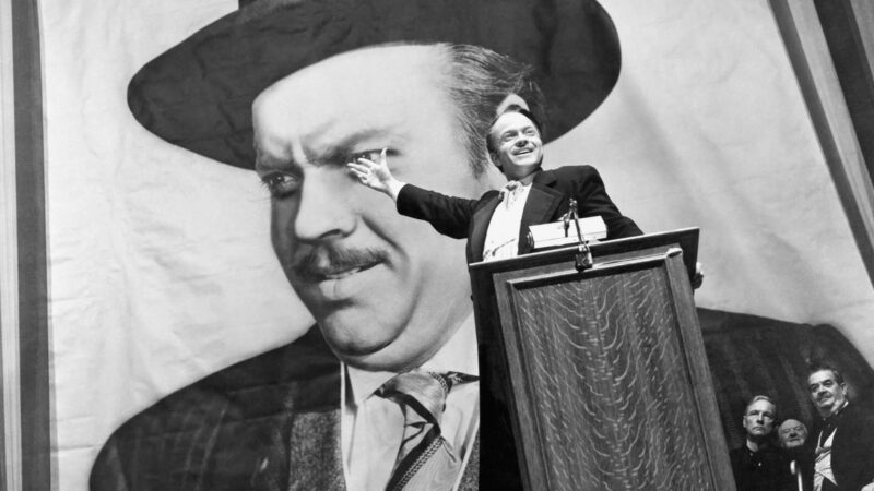 A man stands at a podium giving a speech, smiling and gesturing toward a large portrait of himself behind him. Several people are seated at the side of the stage, looking on. The setting appears formal and dramatic.