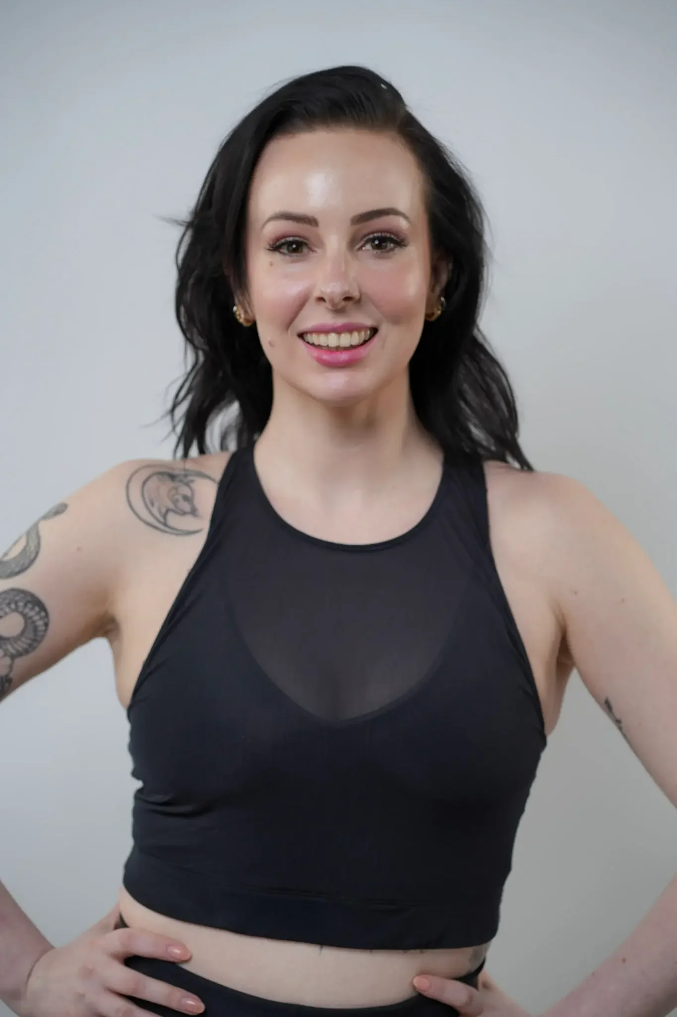 A woman with dark hair, wearing a black sleeveless top, stands against a plain light background. She is smiling and has tattoos on her upper arms. Her hands are placed on her hips.