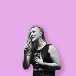 A person with long hair on one side, likely performing at Essex County Nightlife Events, holds a microphone and gestures with their hand on their chest. They wear a sleeveless shirt and earrings, set against a solid light purple background in black and white.