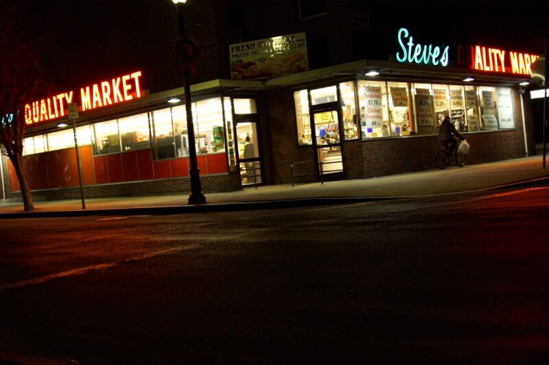 A brick grocery store called “Steves Quality Market” is lit up at night, with glass windows showing shelves and signs inside. A person with a bicycle stands outside near the entrance on a quiet street corner.