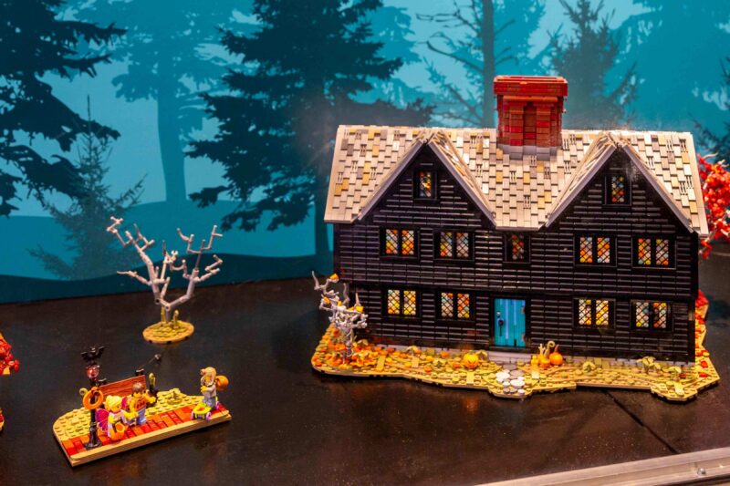 A dark-colored LEGO house with a blue door sits on a surface, surrounded by small trees and figures. Autumn leaves and pumpkins decorate the scene. A painted forest backdrop is visible behind the LEGO display.
