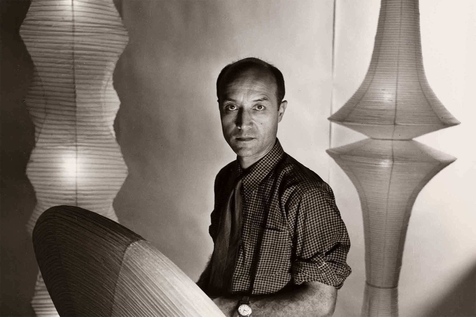 A man with a receding hairline, wearing a checkered shirt and tie, stands indoors between two large, sculptural paper lamps. He faces the camera with a neutral expression. The setting appears to be lit by the lamps, casting soft shadows.