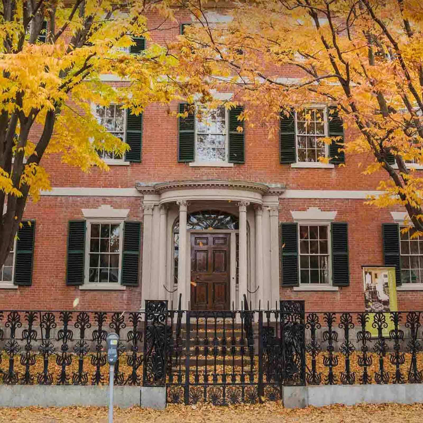 A red brick building with tall windows, black shutters, and a columned entrance sits behind an ornate black iron fence. Yellow autumn leaves cover the ground and trees frame the scene. A parking meter stands at the sidewalk in front.
