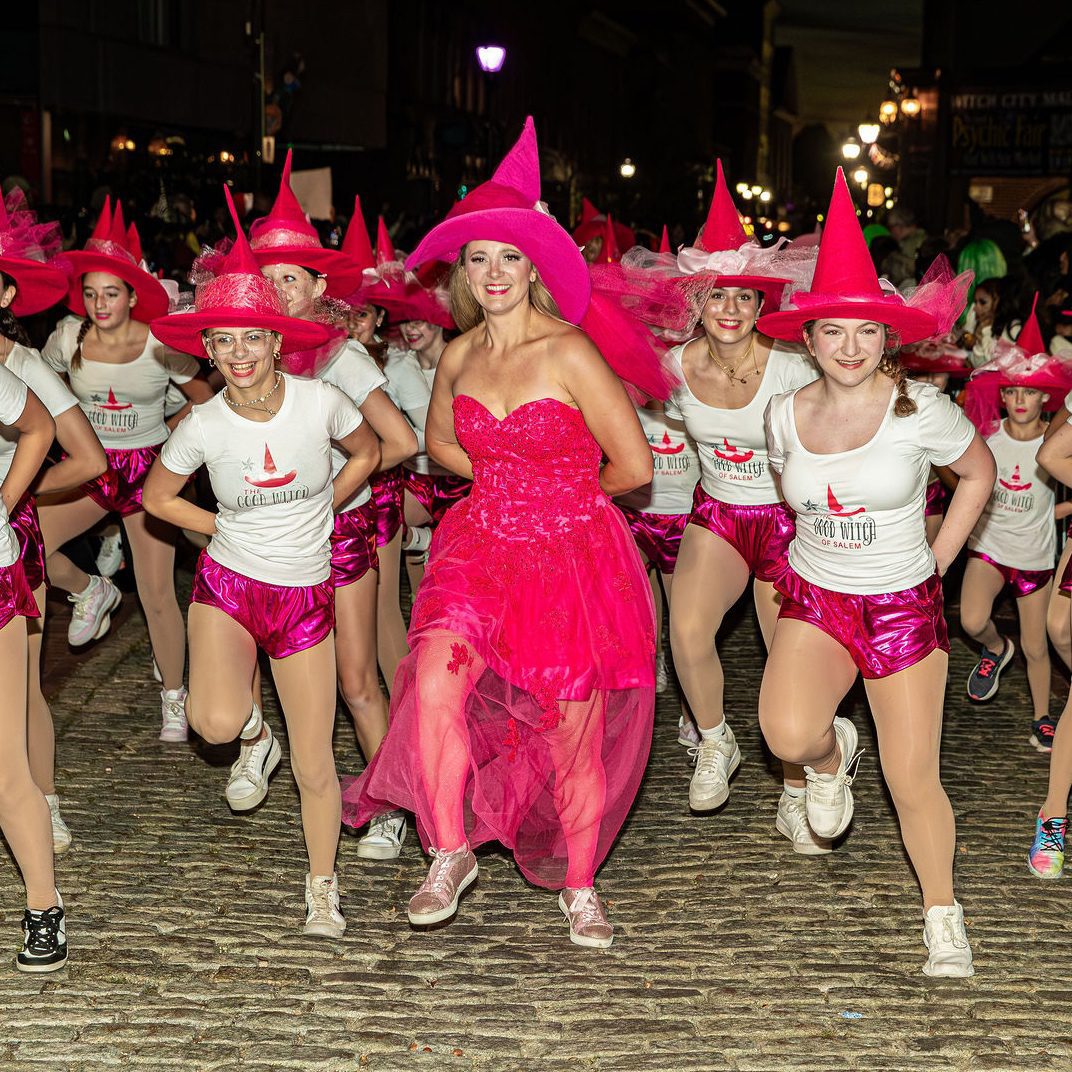 A group of people in white shirts, pink shorts, and pink witch hats dance on a cobblestone street at night. One person in the center wears a bright pink dress and hat. Street lights and buildings are visible in the background.
