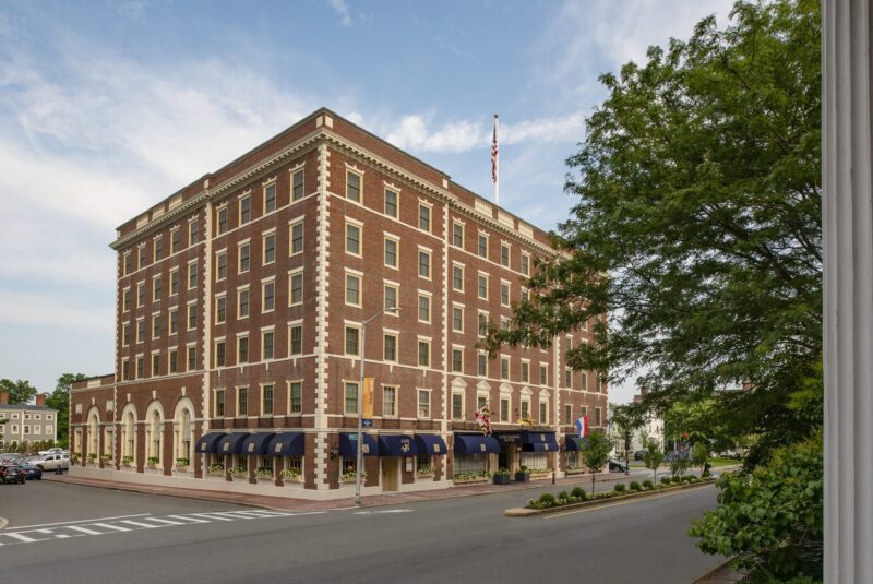 A large, historic brick hotel with five stories features arched windows and blue awnings on the ground floor. An American flag flies atop the building. Trees and parked cars line the streets that surround the hotel.