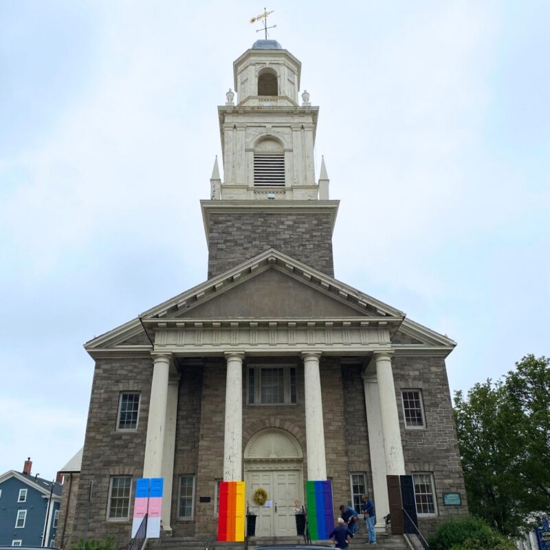 A stone church with white columns and a tall steeple. Six rainbow-colored banners are displayed on the front, and two people are standing near the entrance. The sky is overcast and trees and buildings are visible on either side.
