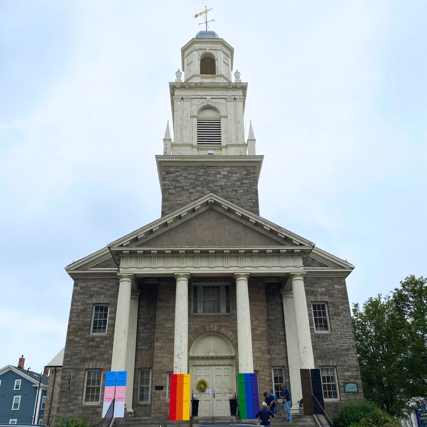 A stone church with white columns and a tall steeple. Six rainbow-colored banners are displayed on the front, and two people are standing near the entrance. The sky is overcast and trees and buildings are visible on either side.