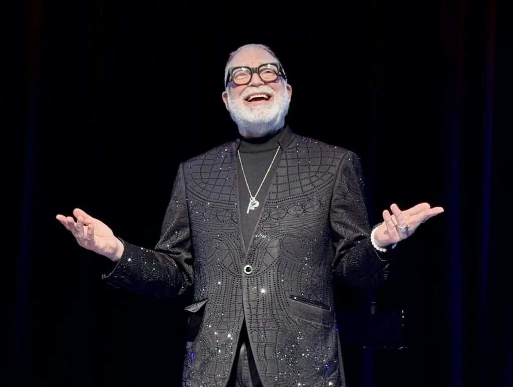An older man with white hair and a beard smiles while raising both hands. He wears glasses, a sparkly black suit, a black turtleneck, and jewelry, standing against a dark background.