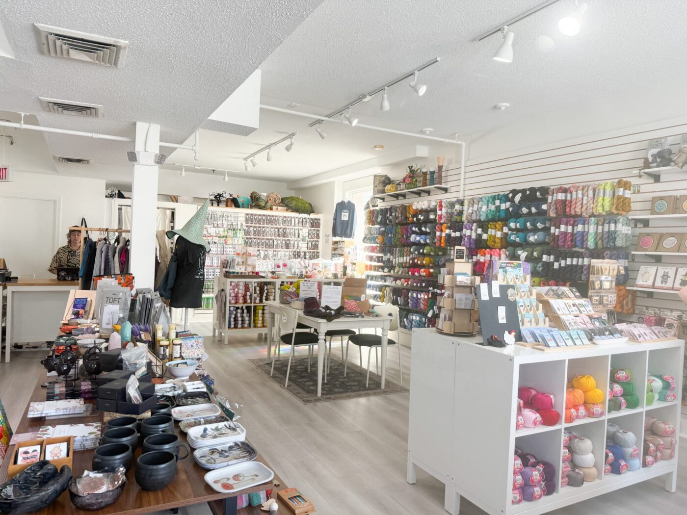 A bright, modern shop interior with shelves of colorful yarn, art supplies, and gifts. A table with yarn sits in the center. Display racks line the walls. A person stands behind the counter in the back left. Natural light from windows brightens the space.