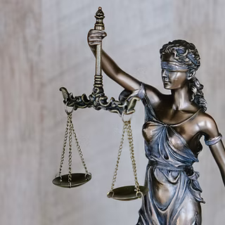 A bronze statue of Lady Justice is shown blindfolded, holding balanced scales in her left hand—a reminder that regular mental health check-ins can help maintain balance amidst life’s uncertainties. The background is blurred and neutral in color.