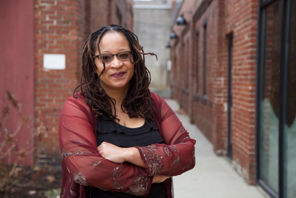 A person with long dreadlocks, glasses, and a sheer red shawl stands outside between two brick buildings, arms crossed, looking at the camera with a slight smile.