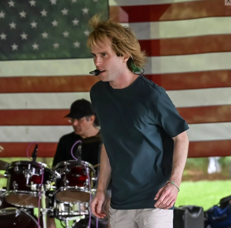 A person with a headset microphone stands in front of an American flag backdrop. A drummer plays behind them. The scene appears to be outdoors.