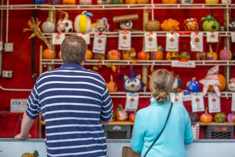 Two people stand in front of a game booth decorated with rows of painted pumpkins and colorful prizes against a red background. The booth has various toys and items displayed on shelves. Both people are facing away from the camera.
