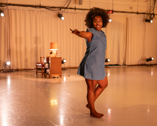 A person stands barefoot on a stage with a polished floor, wearing a short-sleeved denim dress. They are smiling and gesturing with one arm extended. In the background, there is a small table, chair, and lamp with spotlights aimed at the scene.