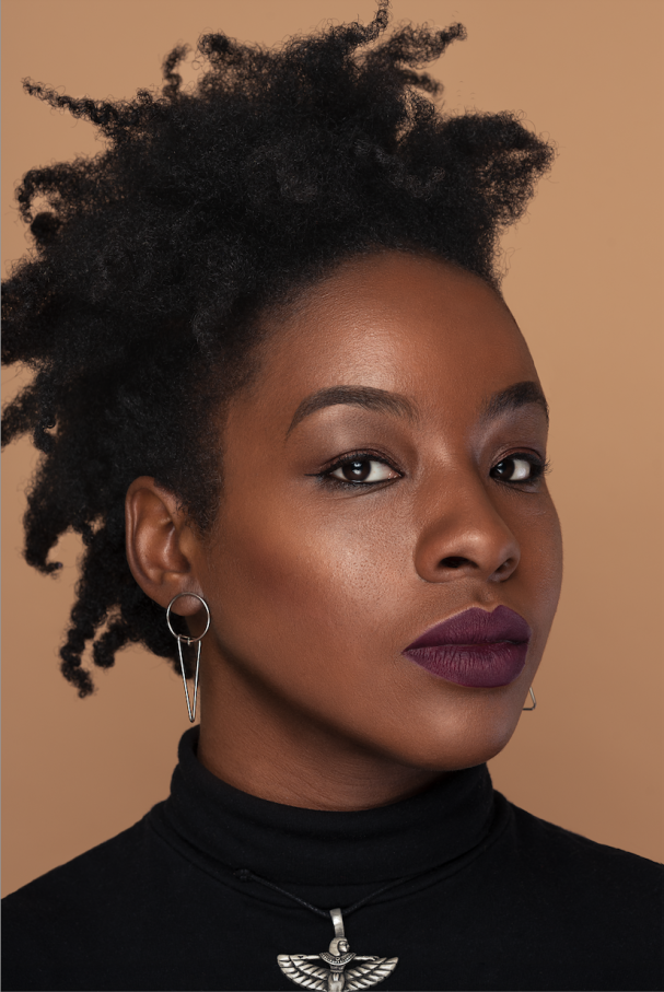 A person with natural, textured black hair and deep plum lipstick looks directly at the camera. They wear a black turtleneck, silver hoop earrings, a geometric pendant, and have a calm, confident expression against a plain beige background.