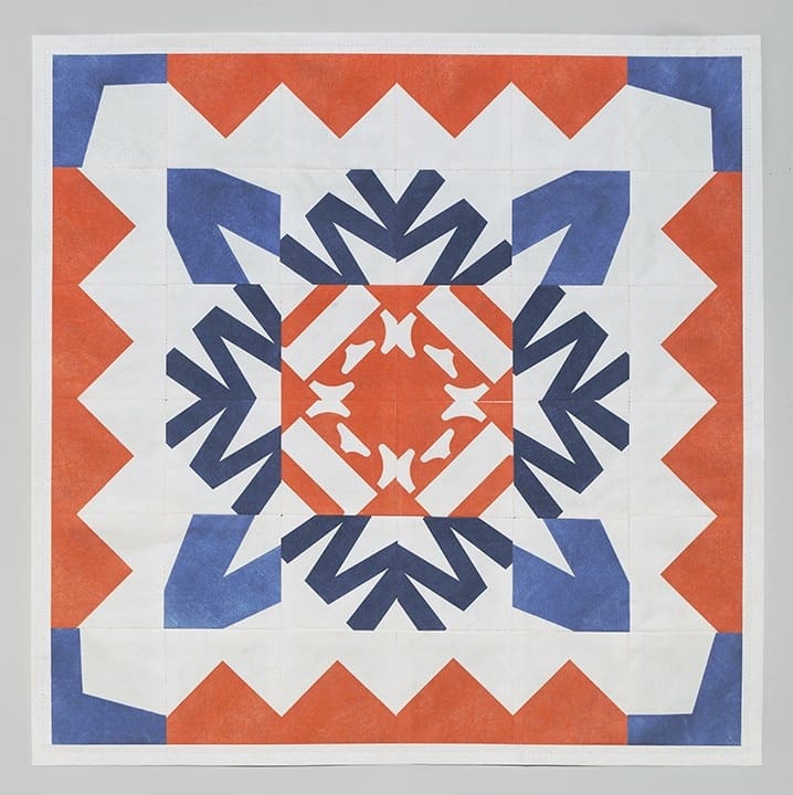 A quilt with geometric patterns in orange, dark blue, and white. The design features symmetrical shapes including triangles and angular forms, arranged around a central square. The overall pattern creates a bold, repeating motif.