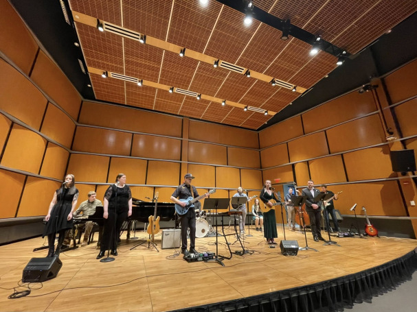 A group of nine musicians stand and sit on a wooden stage in a soundproofed room, playing various instruments including guitars, keyboard, and drums. Microphones and stands are set up, and cables run across the stage floor.
