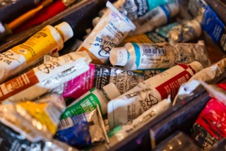 An assortment of paint tubes in varying colors, some squeezed and worn, are scattered in a container. The tubes display different brands and labels, with visible caps and bits of paint residue on several surfaces.