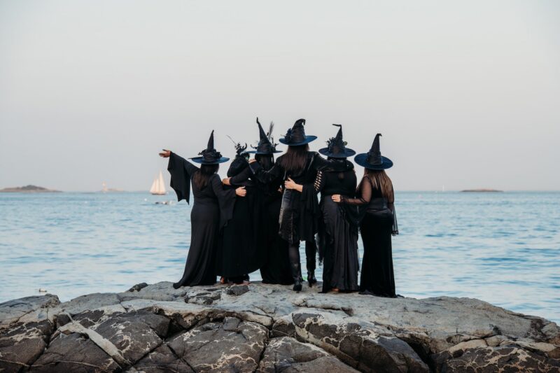 Six people wearing black witch hats and black clothing stand together on rocky ground facing the ocean, with their arms around each other. A white sailboat is visible in the water, and small islands are in the distance under a clear sky.