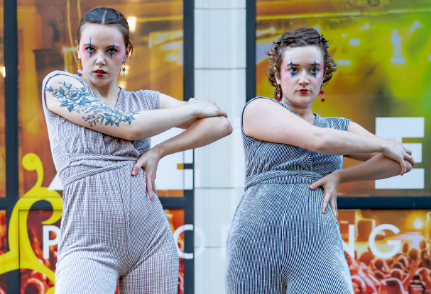 Two people in striped jumpsuits pose with arms crossed in front of a building. Both have bold makeup and earrings. The person on the left has a large arm tattoo. A crowd or event is visible in the blurred background behind them.