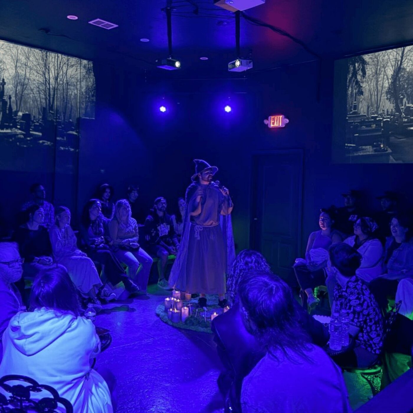 A performer in a witch costume stands on a small stage surrounded by an audience in a dark, blue-lit room. Black-and-white cemetery scenes are projected on the walls. The audience sits on all sides, watching the performer.