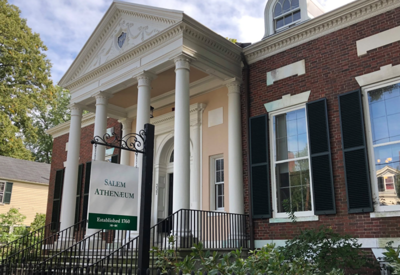 A red brick building with white columns and black shutters, the Salem Athenaeum exudes a convivial craft atmosphere. A black railing lines the steps to the entrance, with lively greenery softening its historic charm.