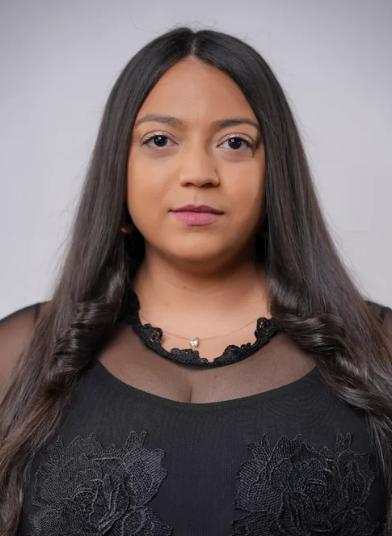 A woman with long, straight dark hair wears a black top with floral lace and a sheer neckline, plus a silver necklace—ready for Hip Hop classes or Beginner Hip Hop—facing the camera with a neutral look against a plain, light gray background.