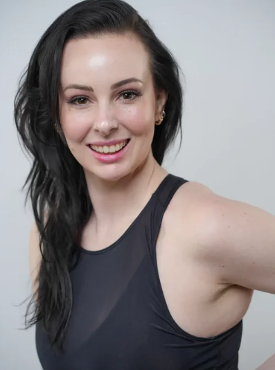A person with long dark hair and a sleeveless black top is smiling at the camera against a plain light background.