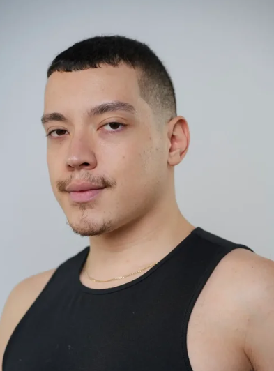 A young man with short dark hair, a trimmed mustache and goatee, wearing a black sleeveless shirt, looks at the camera with a neutral expression. The plain, light background hints at a Heels Level 1 or beginner heels dance class setting.