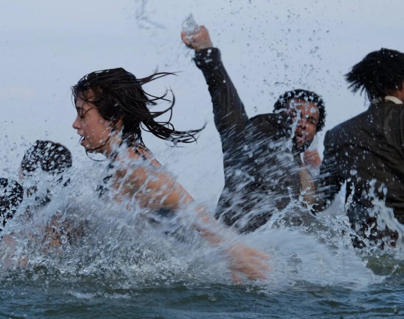 Three people in business attire brave the waves, reminiscent of a RoadShow. One stands in the foreground, another raises an arm behind, ScreenDance energy blending with the blurred background and overcast sky.