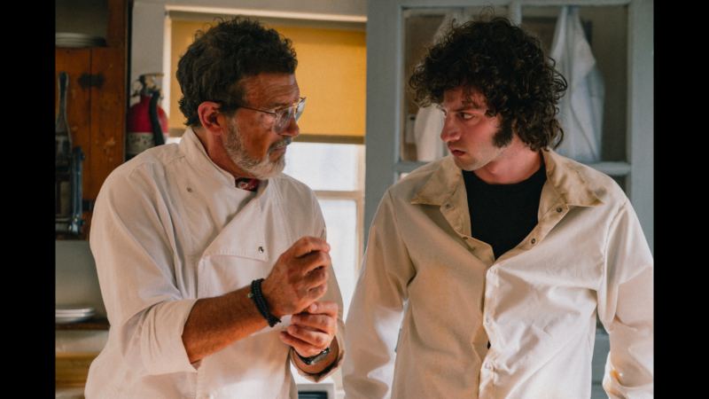Two men in white chef jackets stand in a kitchen. The older man with glasses gestures while speaking, and the younger man with curly hair listens intently. Shelves, dishes, and a window with yellow blinds are visible in the background.