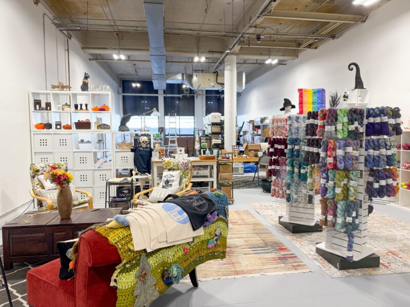 A yarn and craft store interior with colorful yarn displayed on shelves, a seating area with armchairs and a red couch, various home decor items, and large windows at the back letting in natural light.
