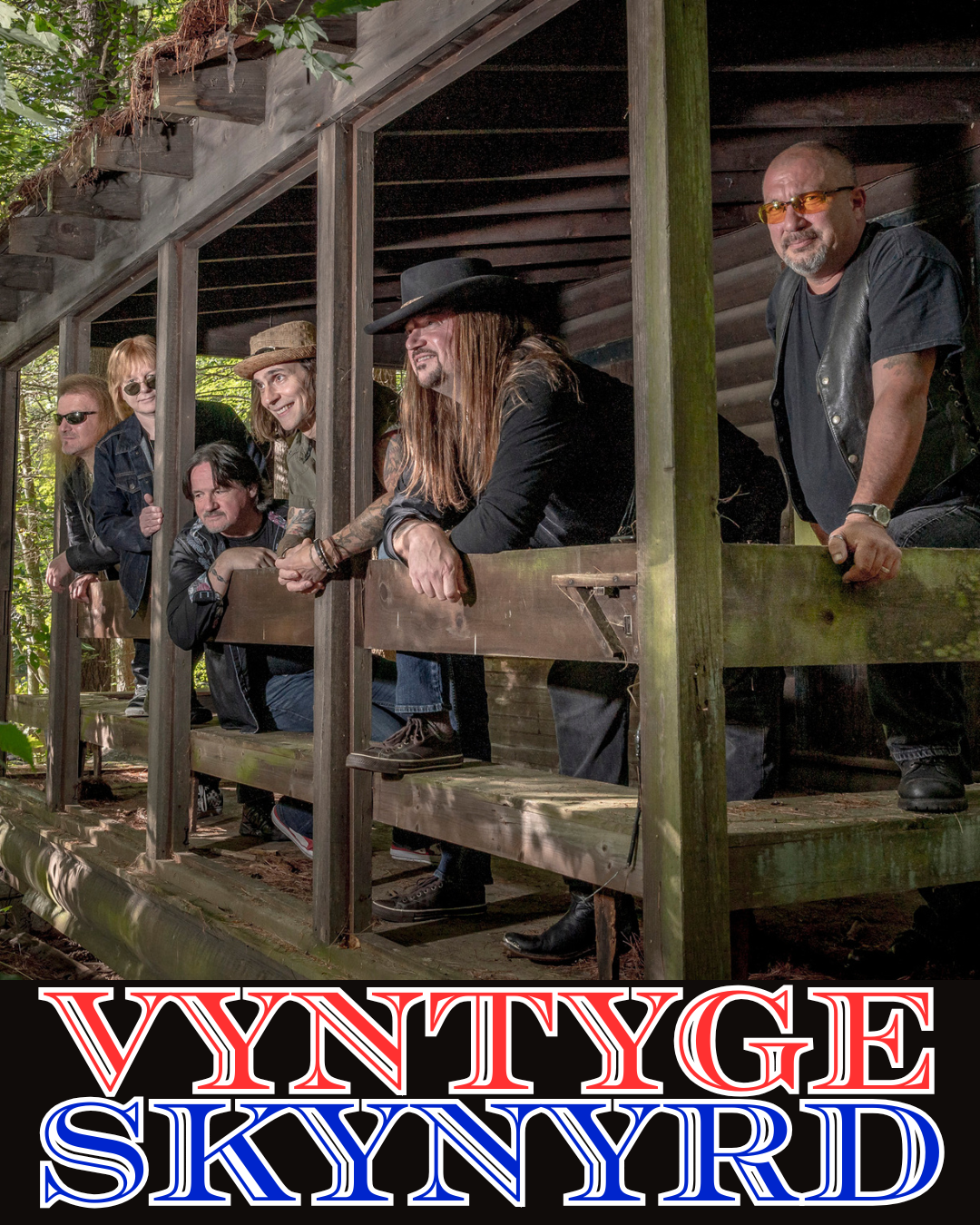 Six men are standing and leaning on the wooden railing of a rustic porch surrounded by trees. They are wearing sunglasses, hats, and casual clothing. At the bottom of the image, large red, white, and blue text reads “VYNTYGE SKYNYRD.”.