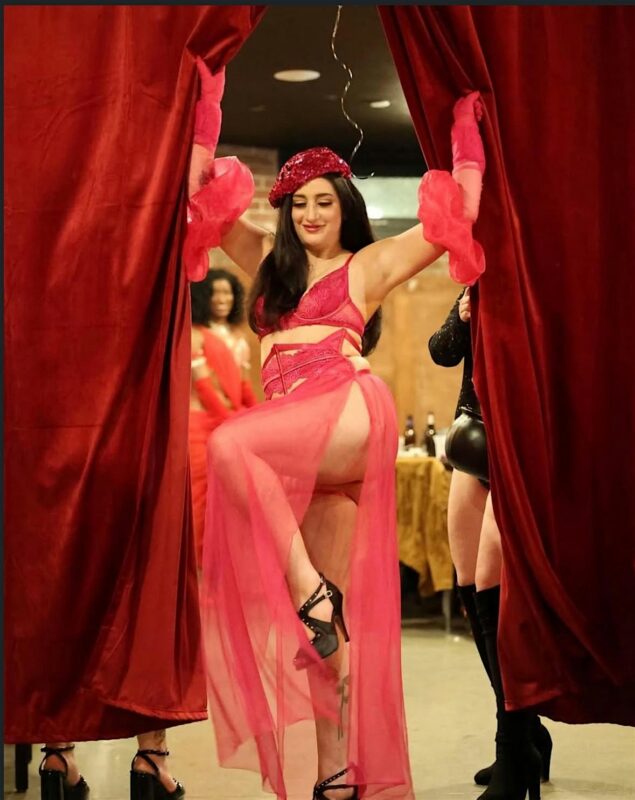 A woman in a bright pink outfit, including a sheer skirt, gloves, and a hat, poses with one leg raised while parting red curtains. Other people stand in the background in a dimly lit room.