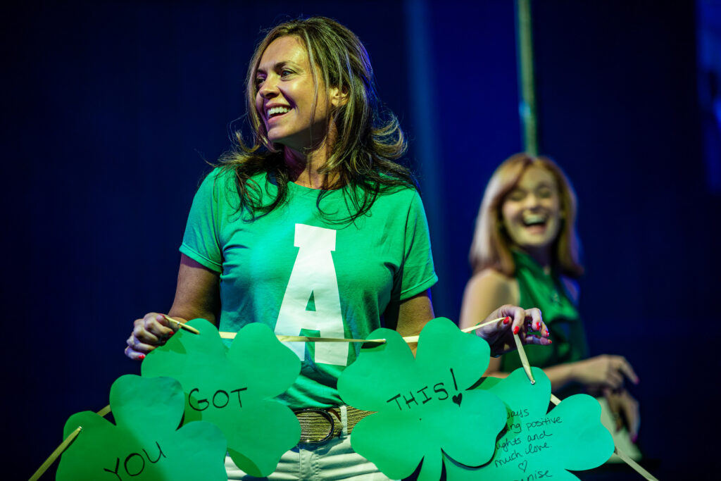A volunteer in a green Patrick's Pack shirt holds up oversized paper shamrocks reading You Got This at A Night for Patrick