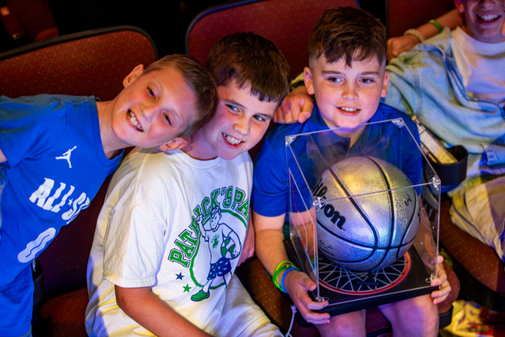 Three boys grin in the audience at North Shore Music Theatre, one holding a signed basketball in a display case, at A Night for Patrick