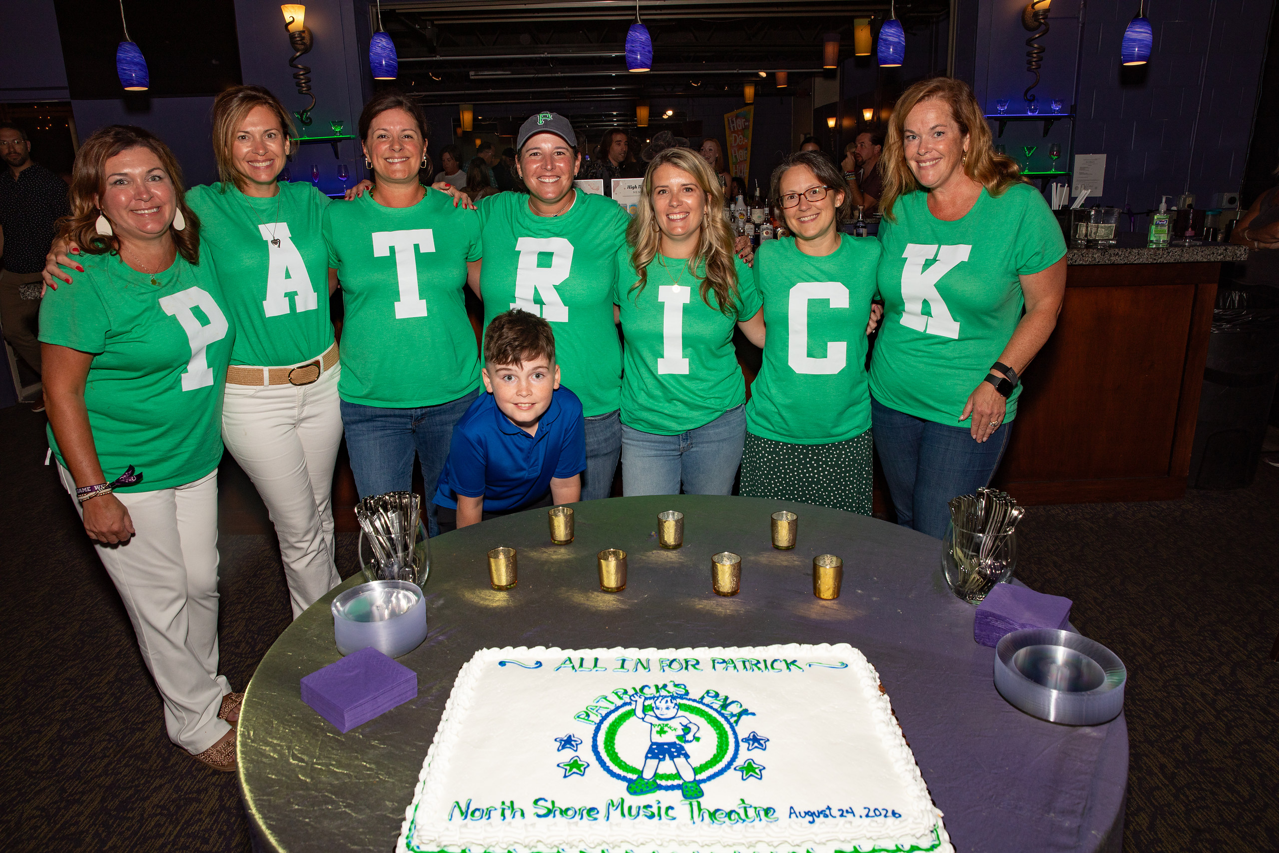 Volunteers in green shirts spelling PATRICK stand behind a sheet cake reading All In for Patrick at North Shore Music Theatre
