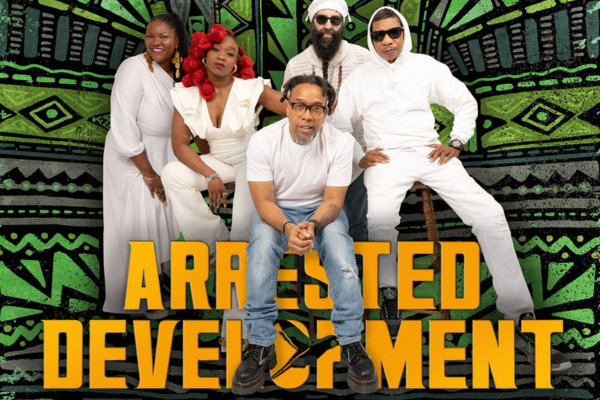Five people pose in front of a green and black patterned backdrop with “ARRESTED DEVELOPMENT” in large yellow letters. Four stand or sit behind one person who is seated at the front. All wear white clothing except the person in front, who wears jeans and a white t-shirt.