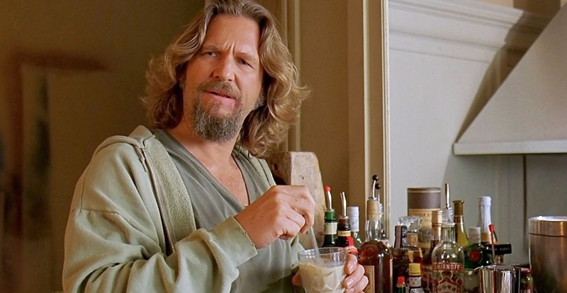 A man with long hair and a beard wearing a gray hoodie stands in a kitchen, holding a glass and a spoon. Bottles of alcohol are visible on the counter next to him. He looks slightly puzzled or confused.