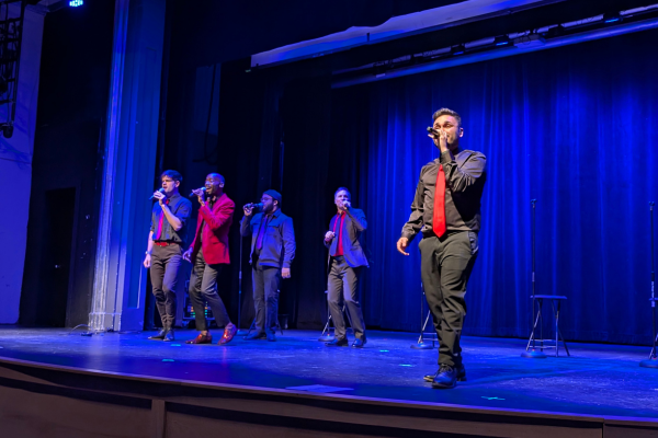 Five men perform on stage with microphones, dressed in suits with red and black accents. The background features dark blue curtains, and the stage is lit with blue lighting. Four men stand to the left, while one man stands at the front right, singing.