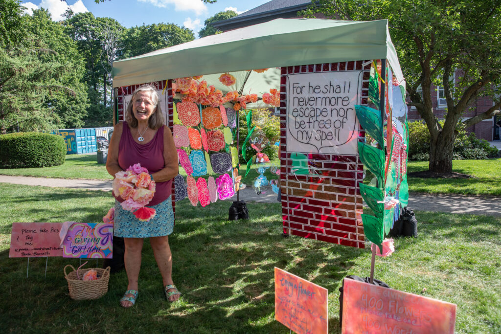 Artist Paula Borsetti laughs beside The Giving Garden, her canopy painted as a brick garden wall and hung with handmade flowers, at the Beverly Garden Party on Beverly Common