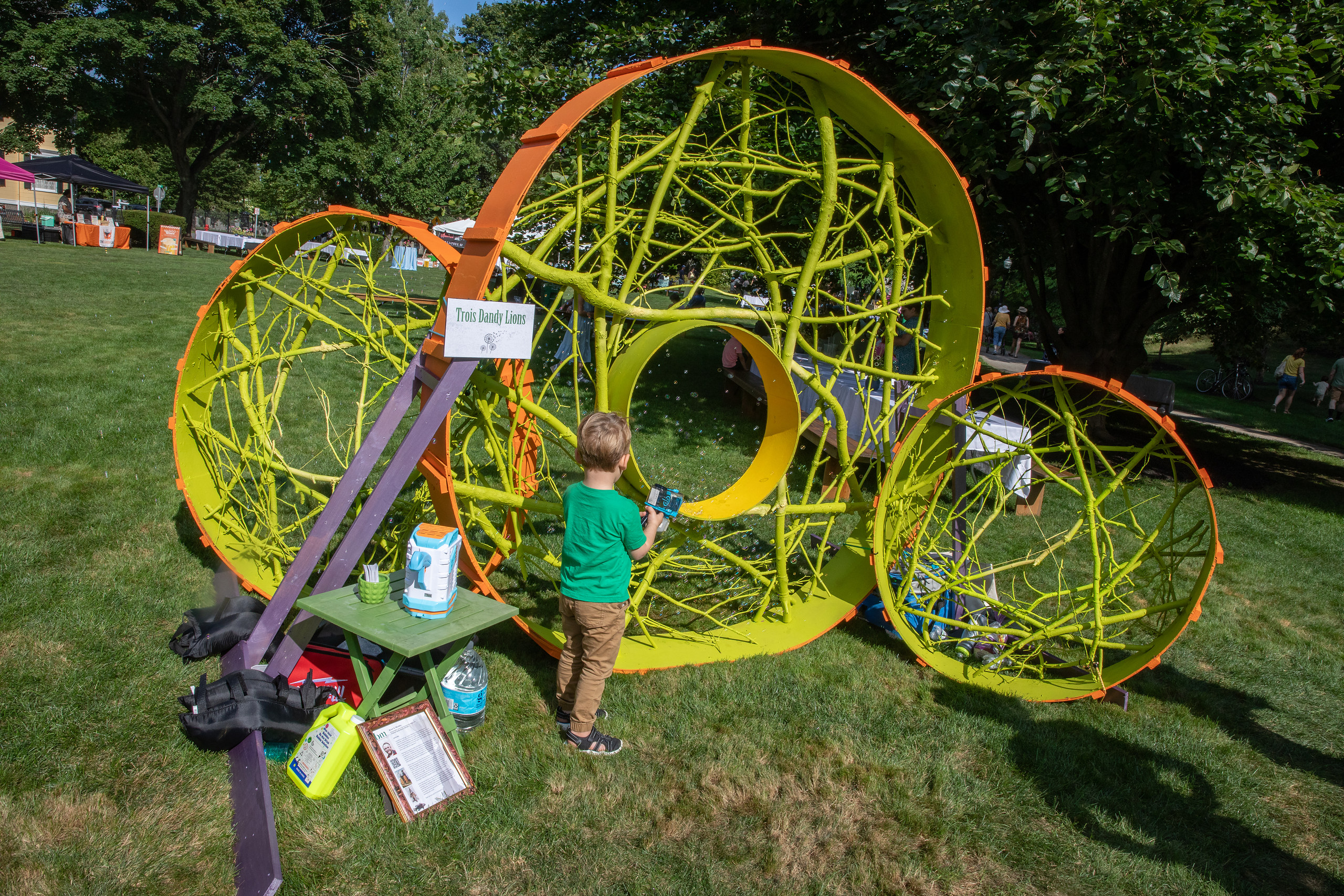 A young boy blows bubbles inside Trois Dandy Lions, a large lime-green sculpture of painted branches formed into three spheres on Beverly Common