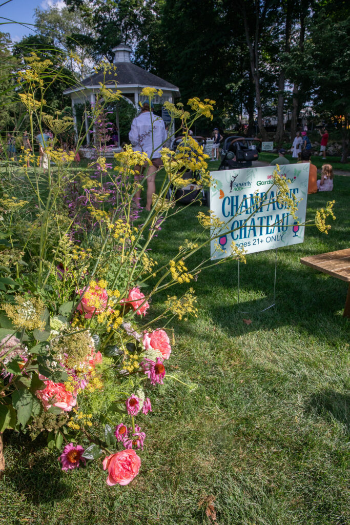 Champagne Chateau sign marked ages 21 and over at Beverly's Garden Party, framed by wildflowers with the Beverly Common gazebo behind it