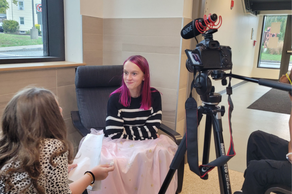 A young person with magenta hair sits in a chair, wearing a black and white striped sweater and pink skirt, facing someone holding papers. A camera with a microphone on a tripod is set up, appearing to record an interview in a bright indoor space.