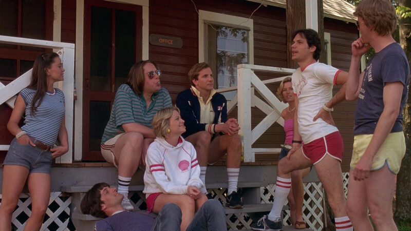 Eight people gather casually outside a brown cabin labeled “Office.” Some sit on the porch, while others stand or sit on the steps. They wear casual summer clothes like shorts and t-shirts, with trees and white porch railings visible in the background.